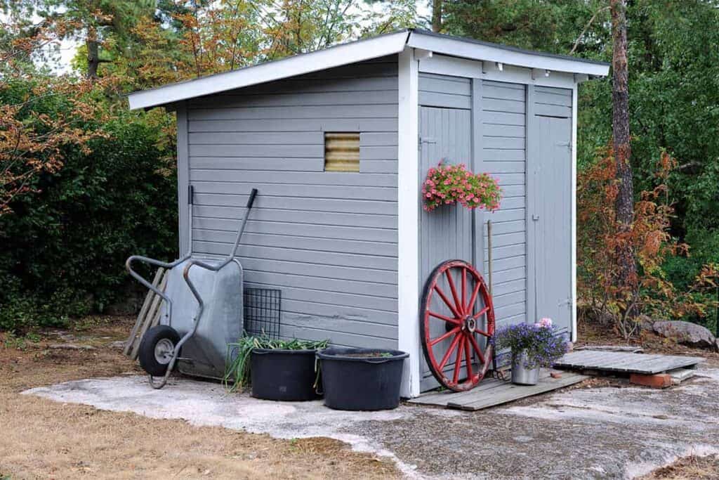 A blue-gray garden shed with a slope roof sits on the edge of a tree line. Outside are flower boxes, a red wagon wheel, and a wheelbarrow.