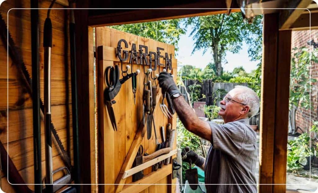 A man hangs garden tools on the back of a garden shed door.