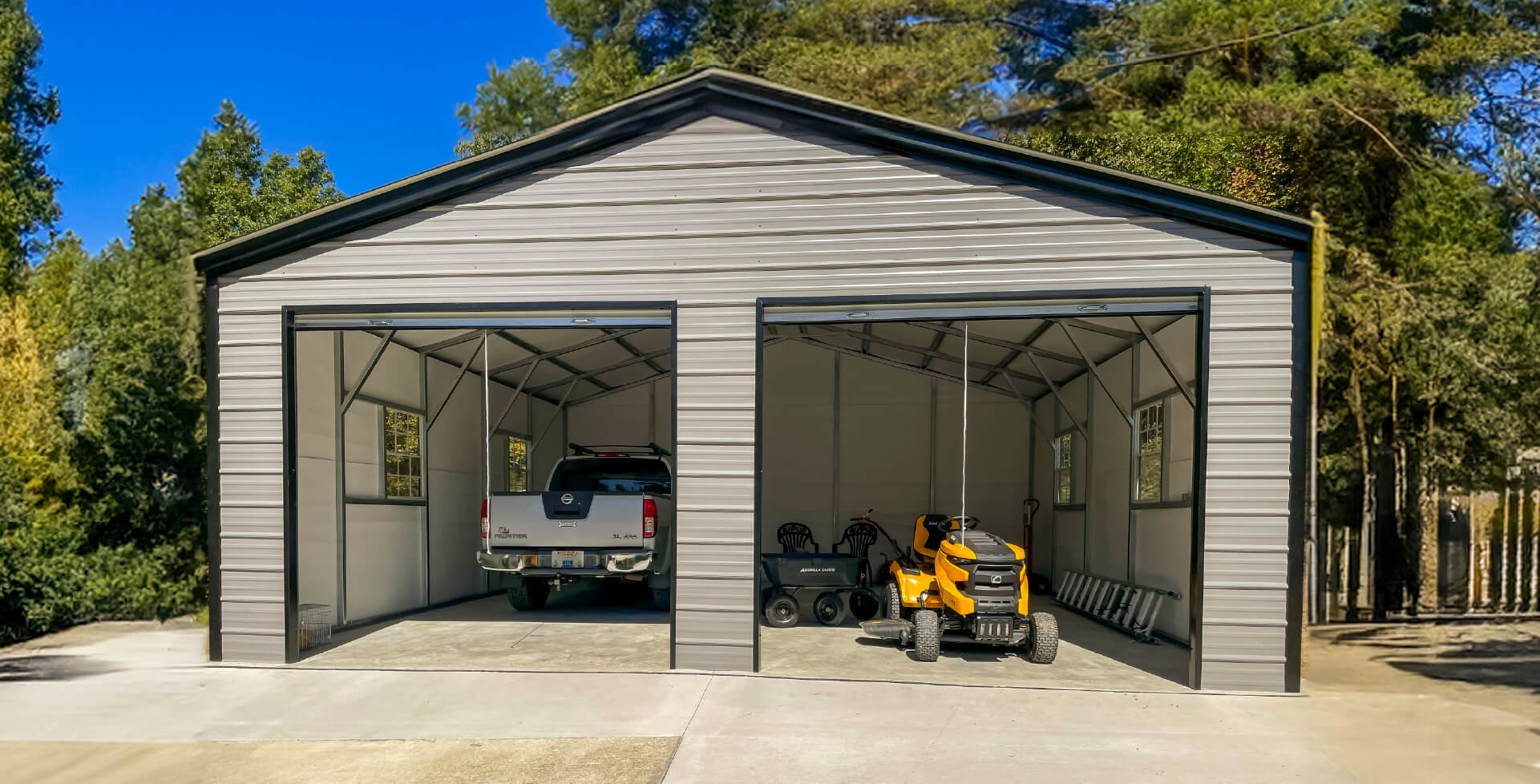A tan two-car detached metal garage with two open bay doors, holding a pickup truck and a yellow lawn tractor, set among trees.