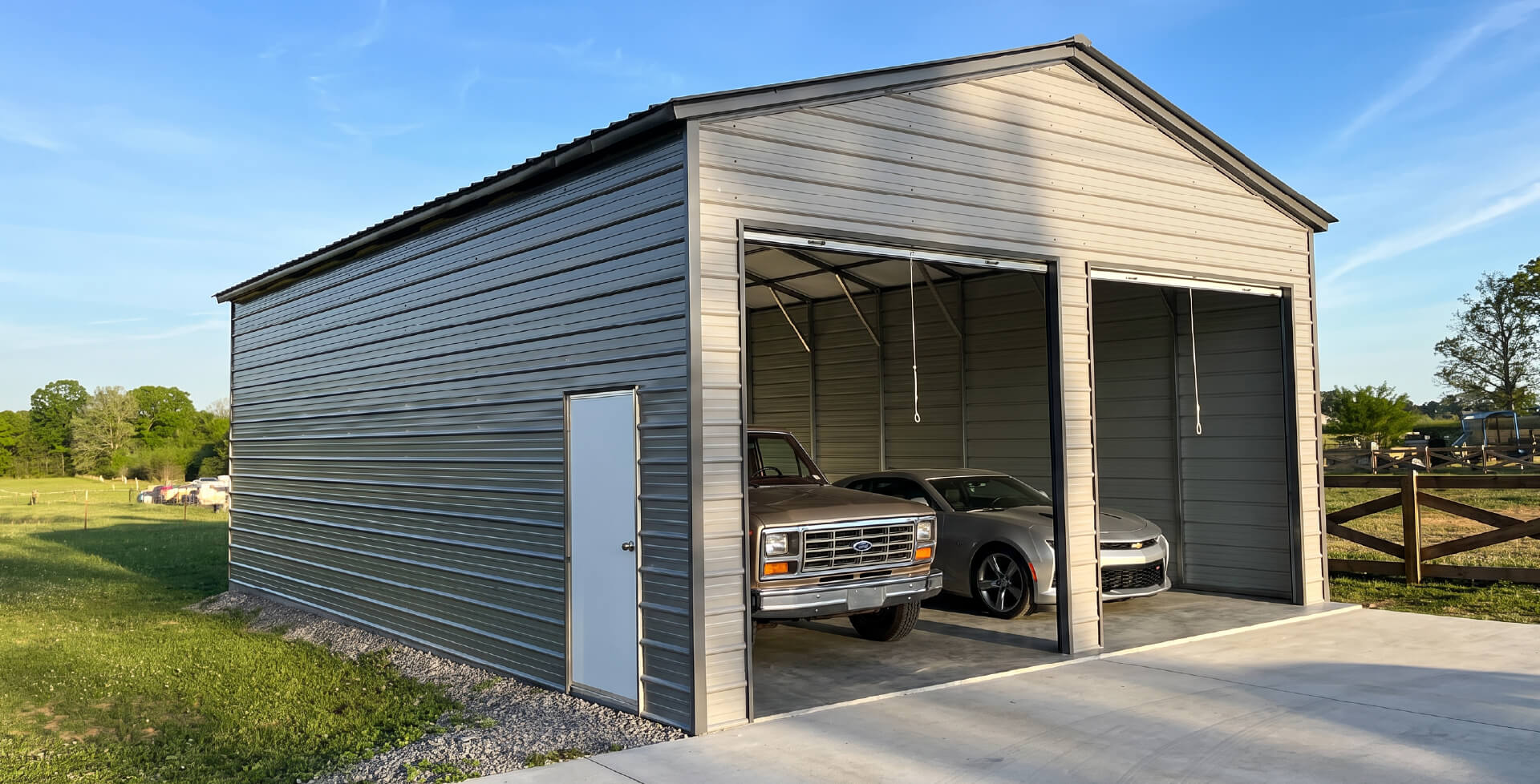 A tan two-car detached metal garage with two open bay doors, holding a pickup truck and a yellow lawn tractor, set among trees.