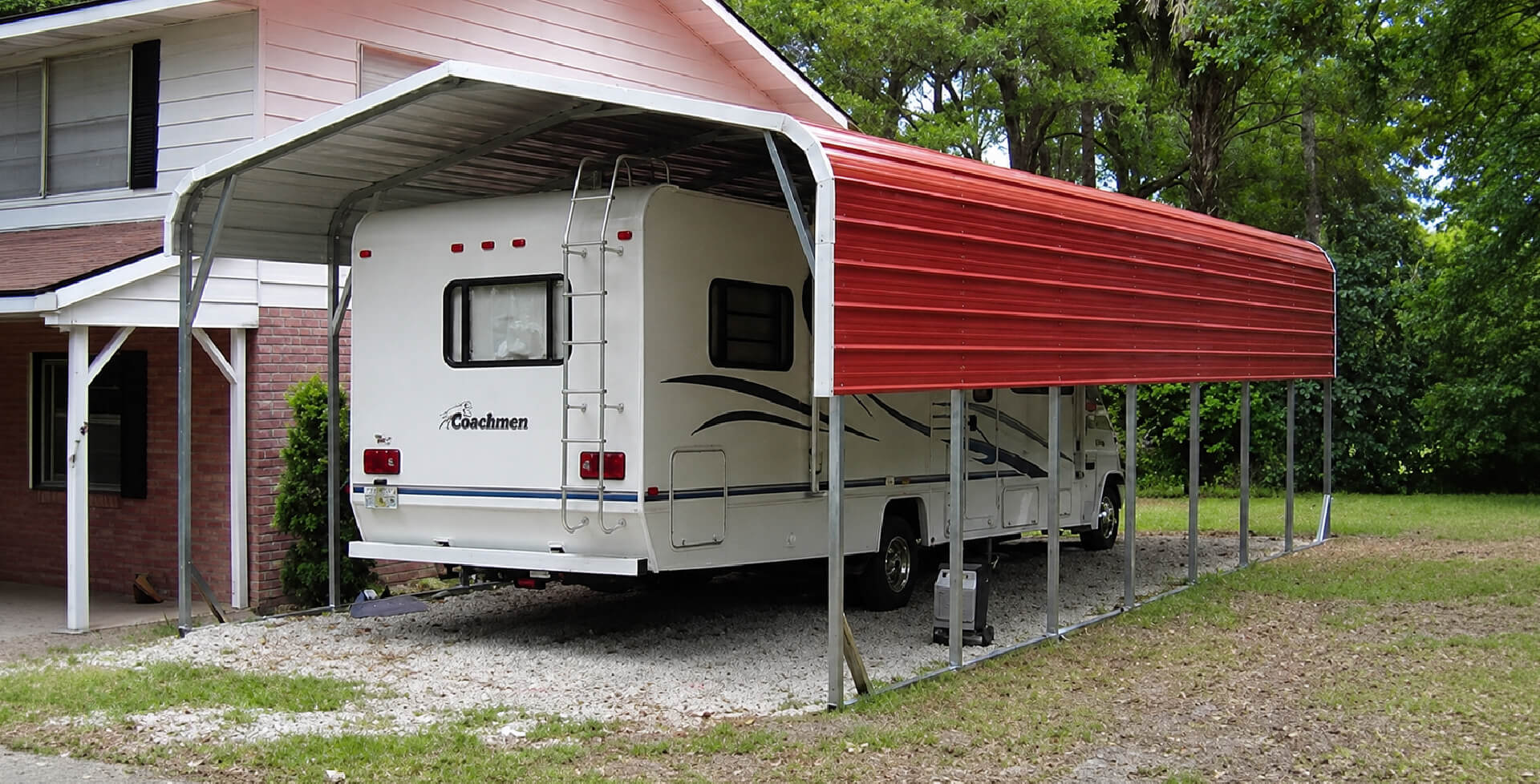 A tall open-sided metal RV carport shelter covering a large travel trailer, with a small storage shed and trees in a wooded yard.