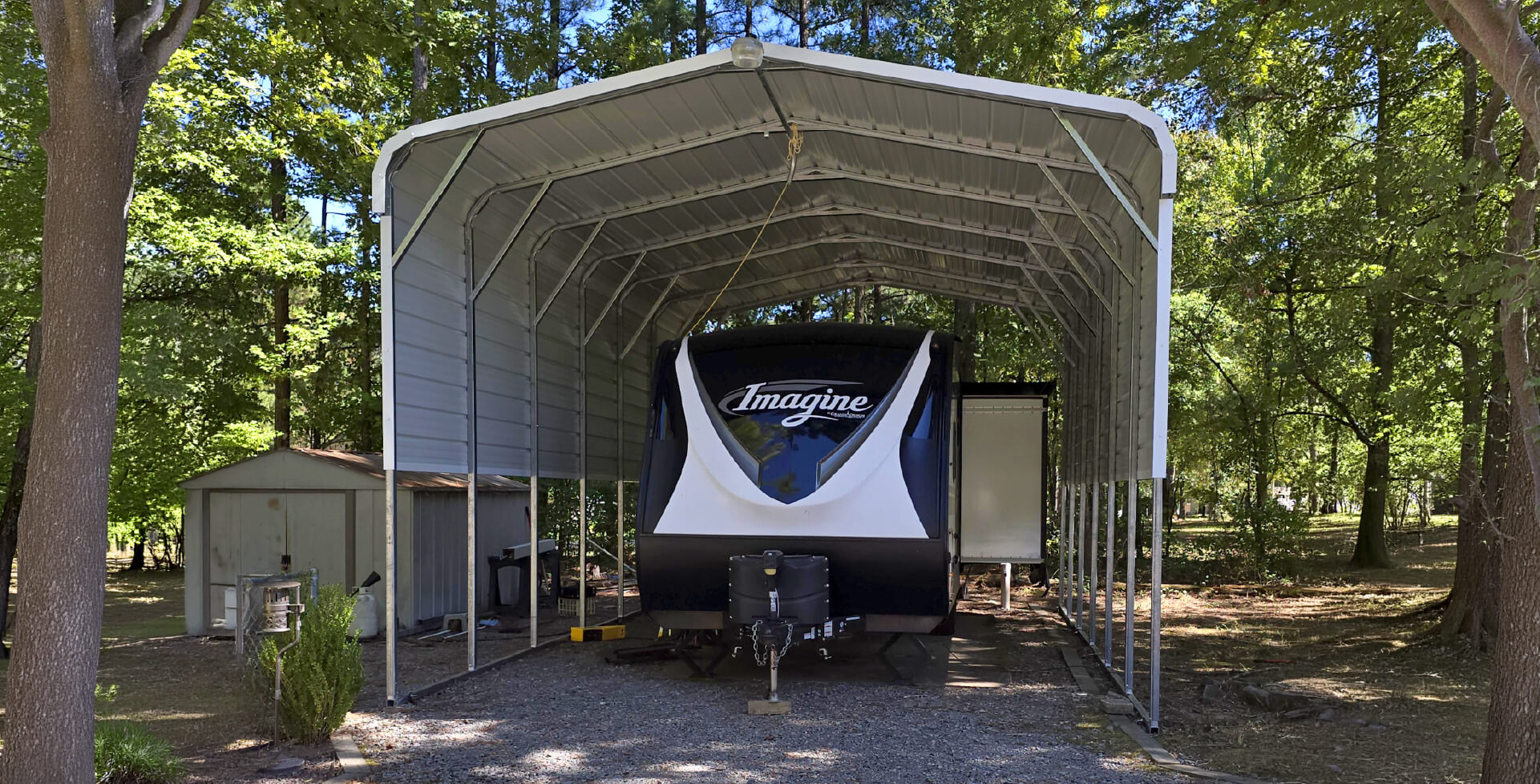 A tall open-sided metal RV carport shelter covering a large travel trailer, with a small storage shed and trees in a wooded yard.