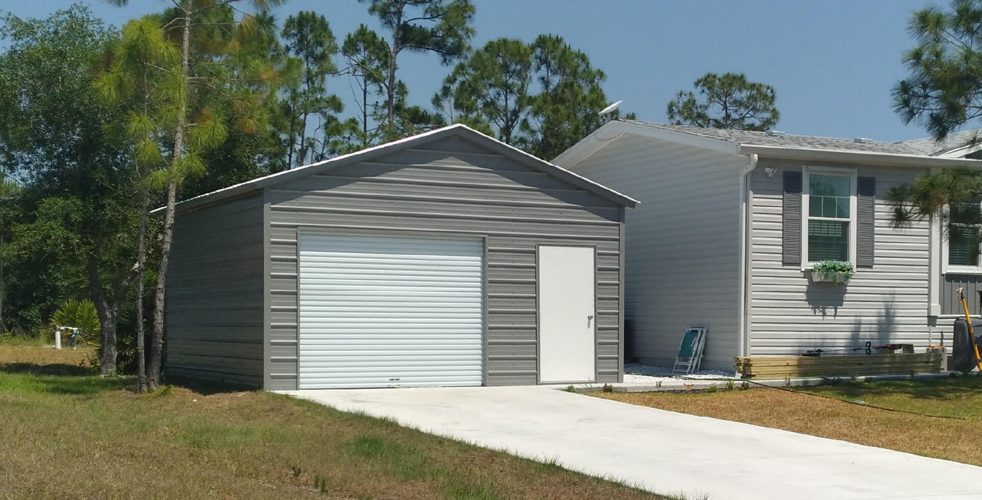 Detached gray metal garage with white roll-up door and side entry beside a residential home