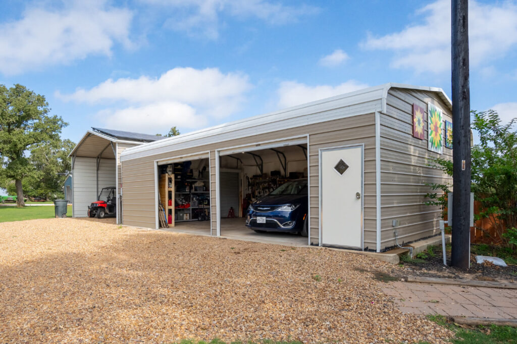 Beige two-car metal garage with open roll-up doors revealing a parked car and an ATV inside, set on a gravel driveway under a blue sky.