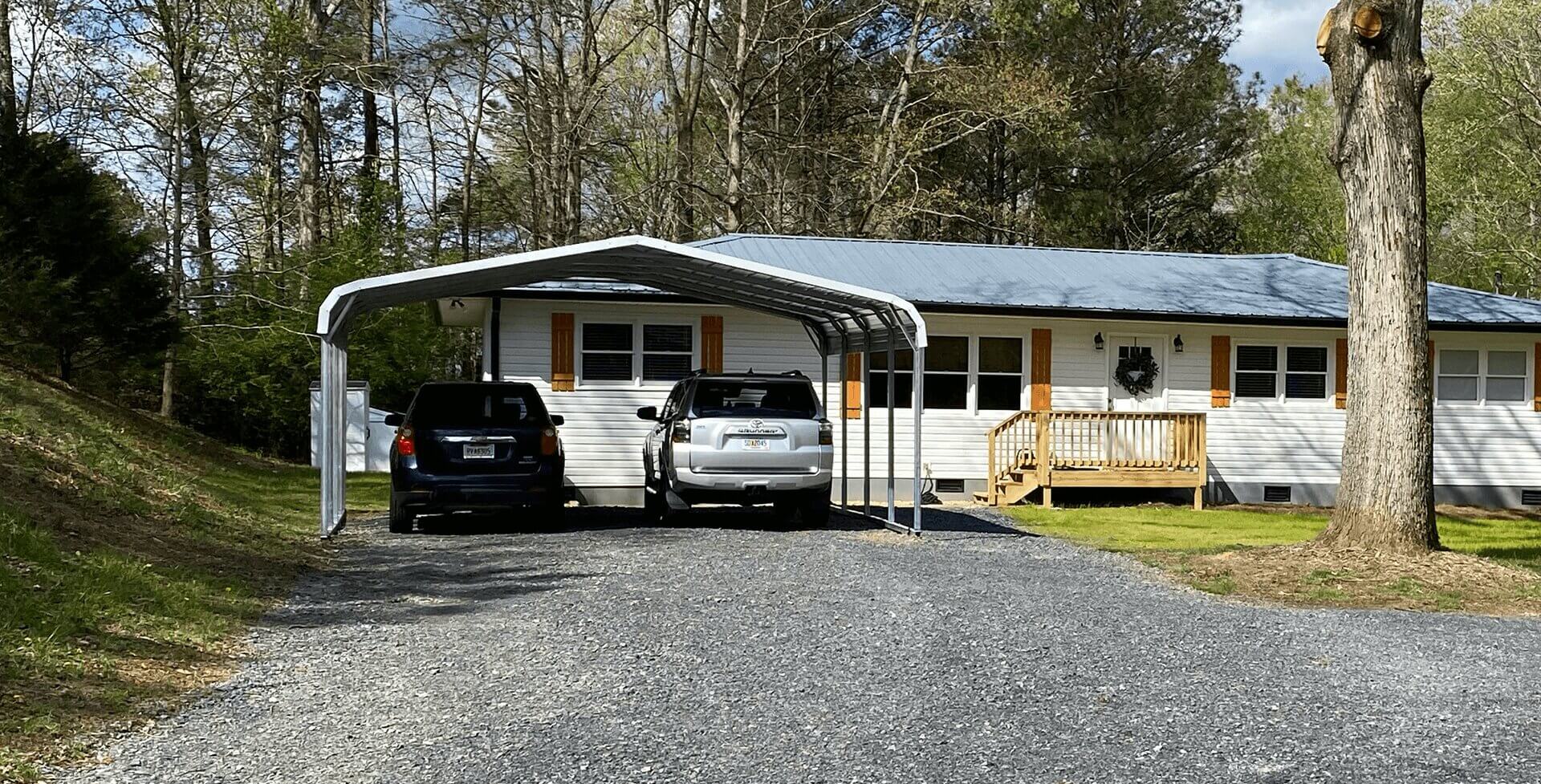 A gravel driveway leads to a metal double carport attached to a single-story white house with wood shutters. Two vehicles—a black SUV and a silver SUV—are parked under the carport.
