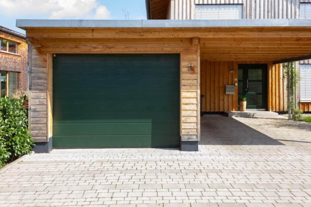 A garage-carport hybrid is at the end of a driveway. The garage is build with tan bricks and has a green door.