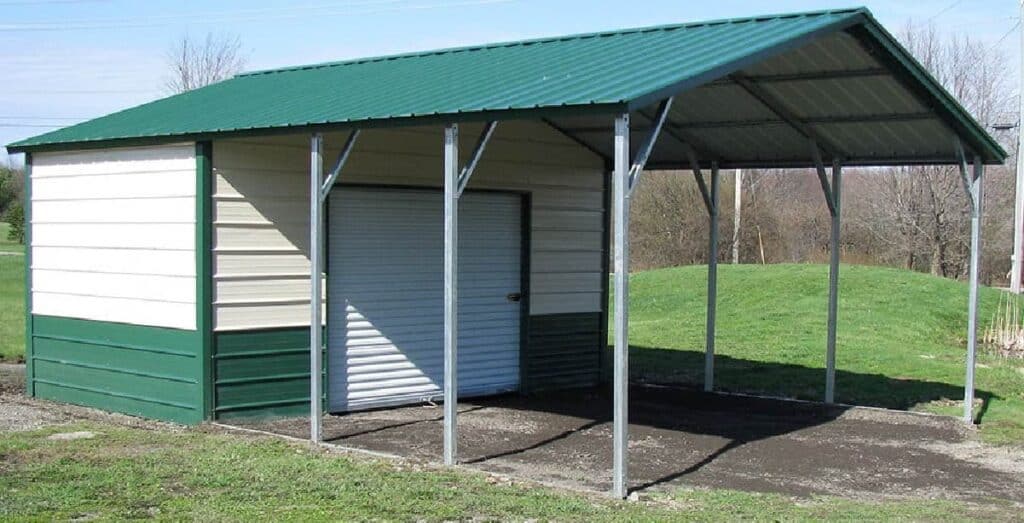 A metal garage with green trim and a white garage door has a green carport extension from its metal roof. The garage is in a grassy field with trees on the horizon.