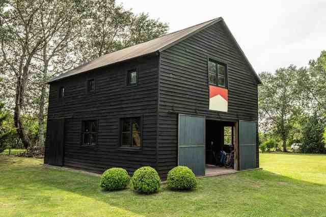 A black, two-story barndominium sits on a grassy lawn with trees in the background. The barn door is open and a red and white flag hangs over the door.