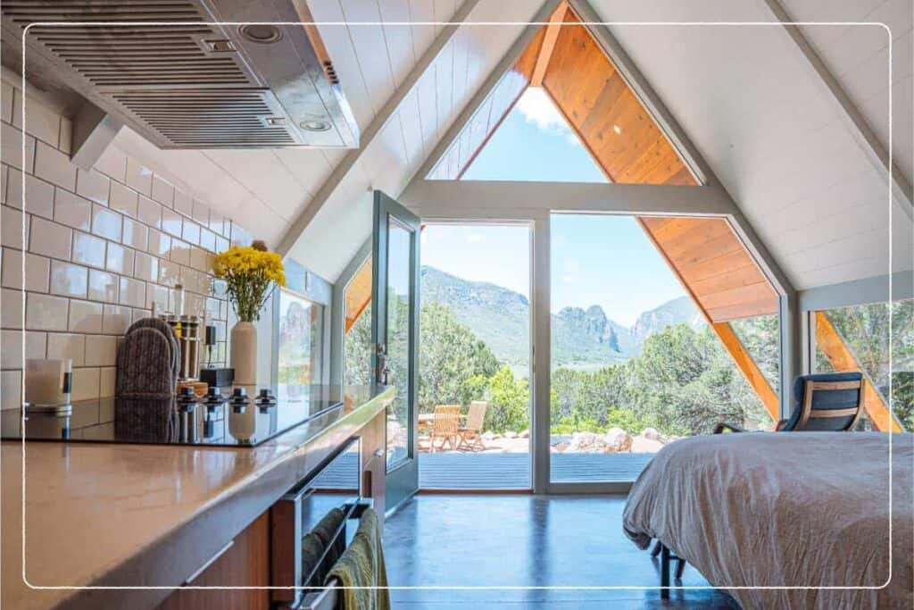 An A-frame home shows a glass wall with a view of trees and mountains.