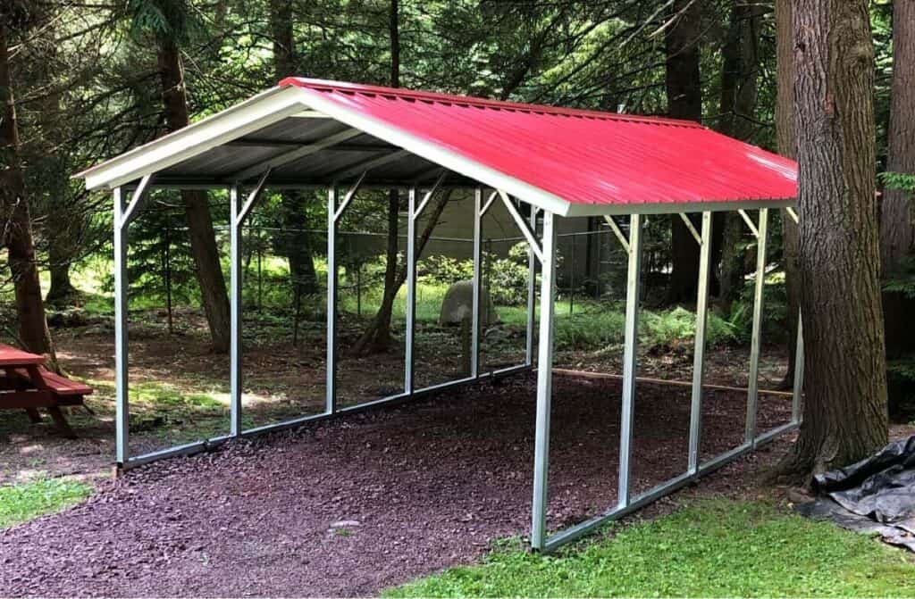 A white carport with a red roof sits in a wooded landscape among the trees.
