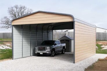 A metal carport shows a gray GMC pickup truck underneath parked on a gravel driveway. The carport is painted beige.
