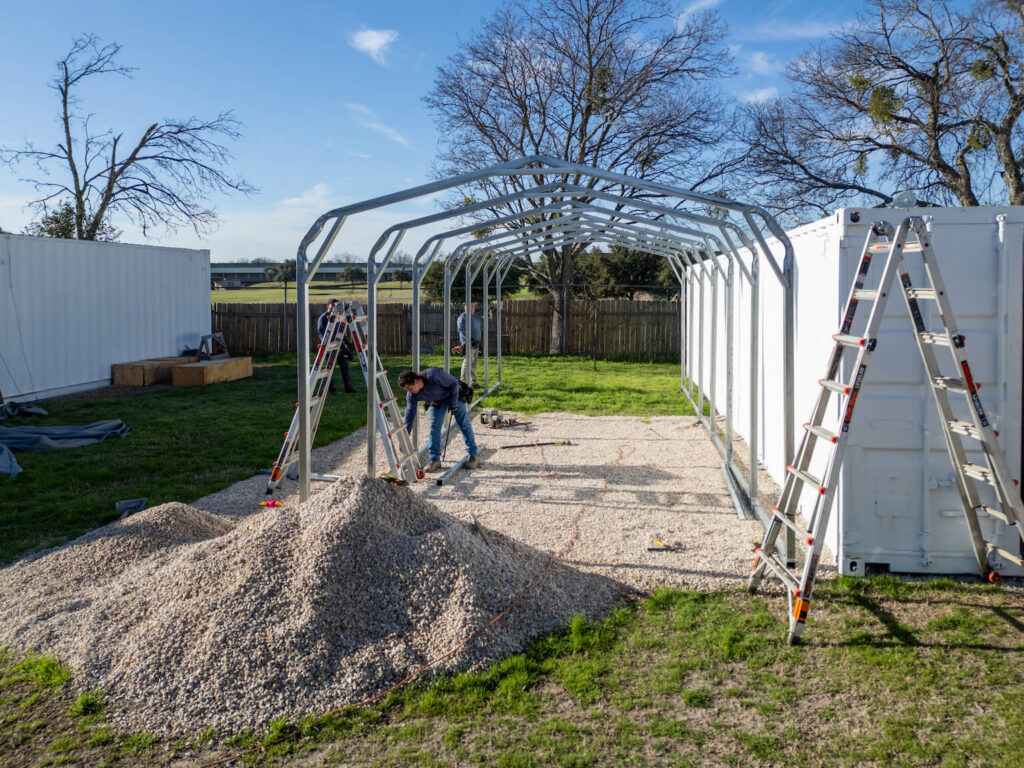 Workers assembling a steel garage frame on a prepared gravel foundation.