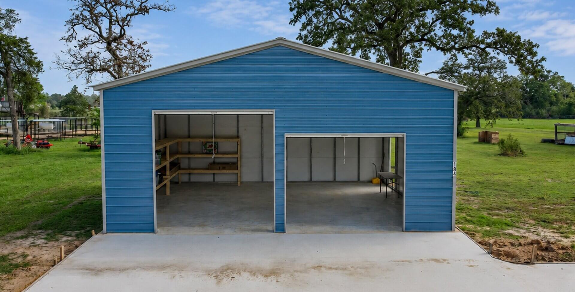 Blue metal garage building with two open bay doors and a gray roof, set on a concrete pad surrounded by grass and trees.