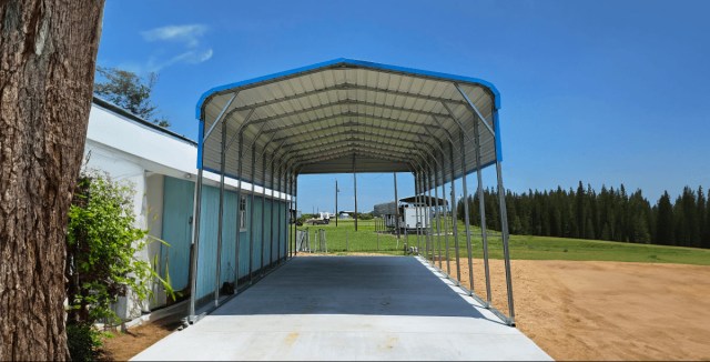 Metal carport with blue trim on a concrete pad beside a building, under a clear sky