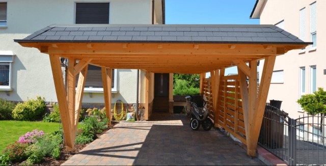 Wooden carport with a shingled roof, attached to a house and featuring a side privacy screen