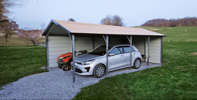 Metal carport with a tan roof sheltering a silver car and a lawn tractor on a gravel base