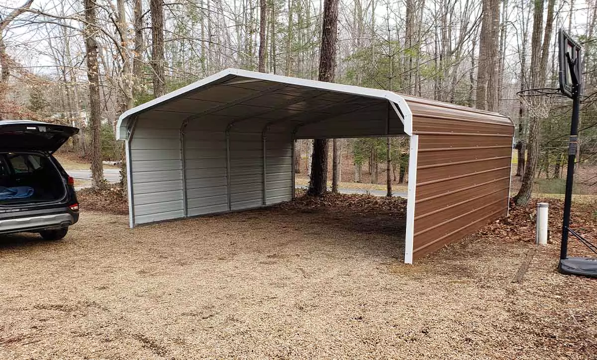 A brown carport with covered sides has two openings connecting a gravel drive with the road. 