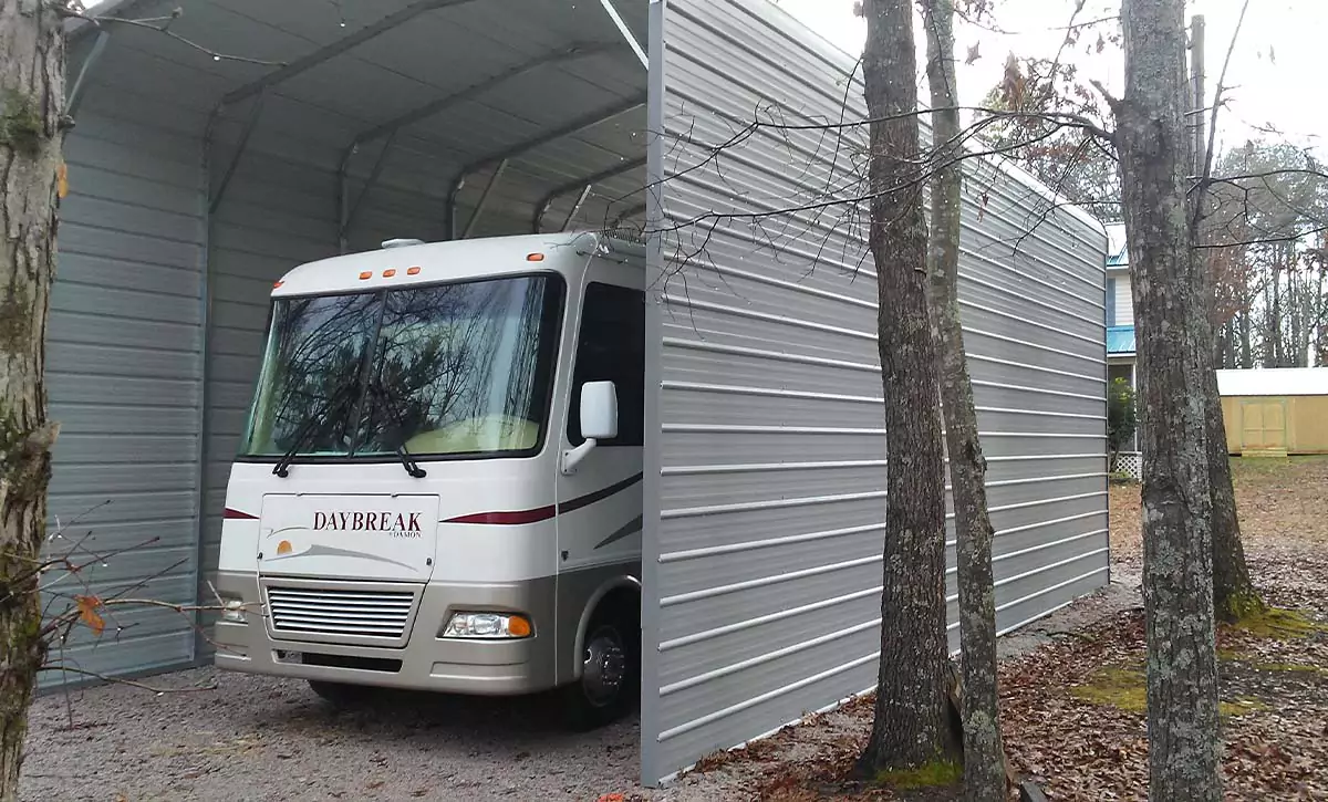 An RV is parked under a metal carport with side walls in a backyard. 