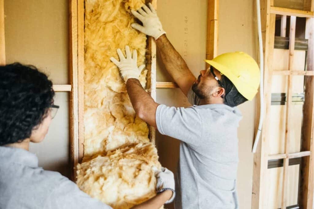 A man installs fiberglass insulation between studs of a garage frame.