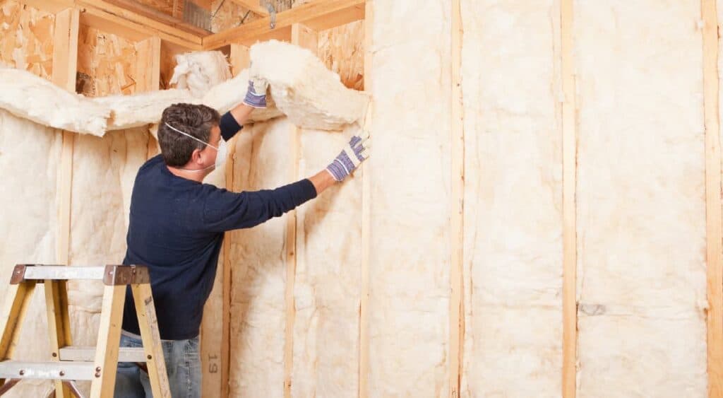 A man wearing safety equipment pushes fiberglass batts between garage studs. 