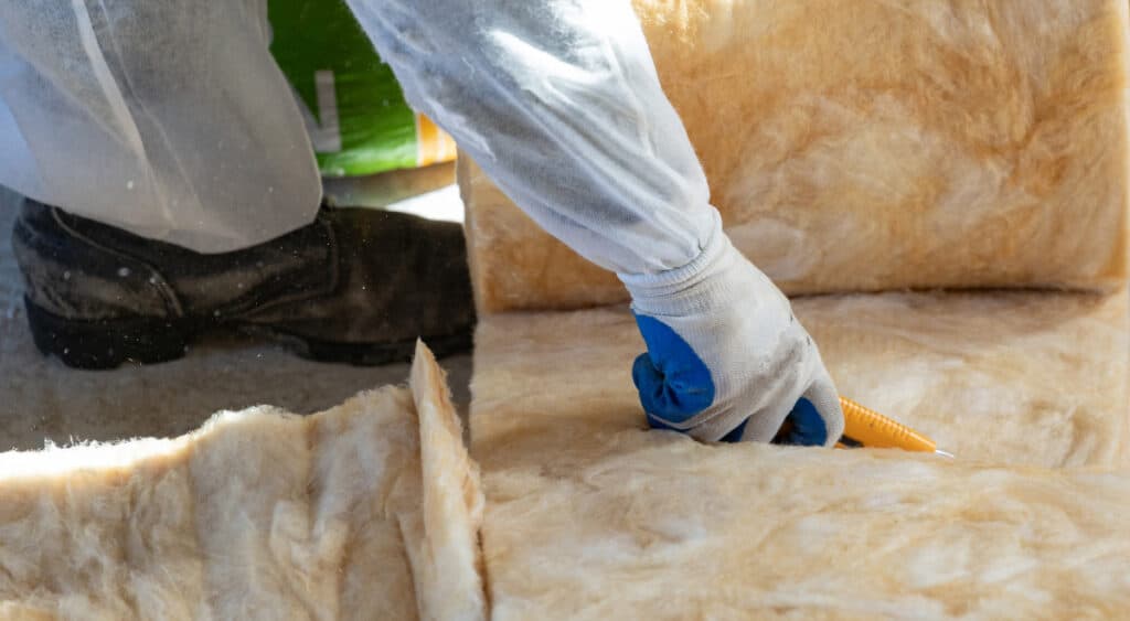 Close up photo of a gloved hand cutting fiberglass insulation with a utility knife.