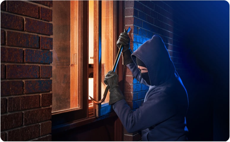 A burglar dressed in a hooded sweatshirt, mask, and gloves trying to break in through a window using a crowbar
