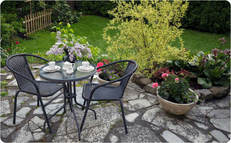 A small patio table with tea setting and chairs on a stone patio surrounded by a grass lawn, potted flowers, and garden plants.
