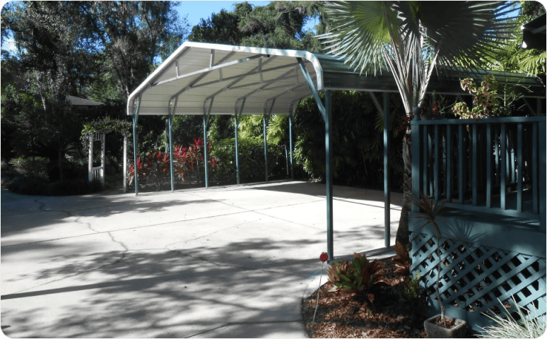 A metal carport surrounded by landscaping including a garden, shrubs, and trees.