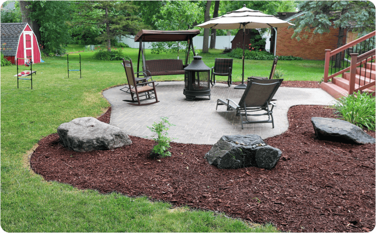 Landscaping featuring lounge furniture on a stone patio surrounded by mulch, rocks, plants, a grass lawn, and trees.