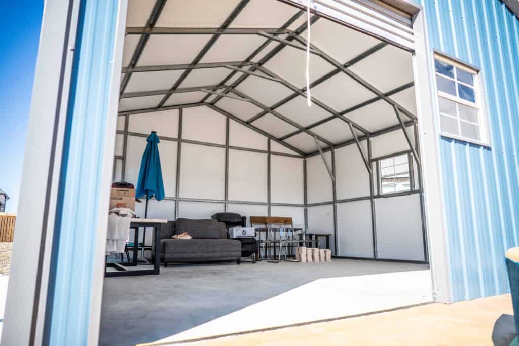 Interior view of a blue metal garage from Alan's Factory Outlet with an open garage door, featuring a sofa, a blue umbrella, a table, and storage boxes.
