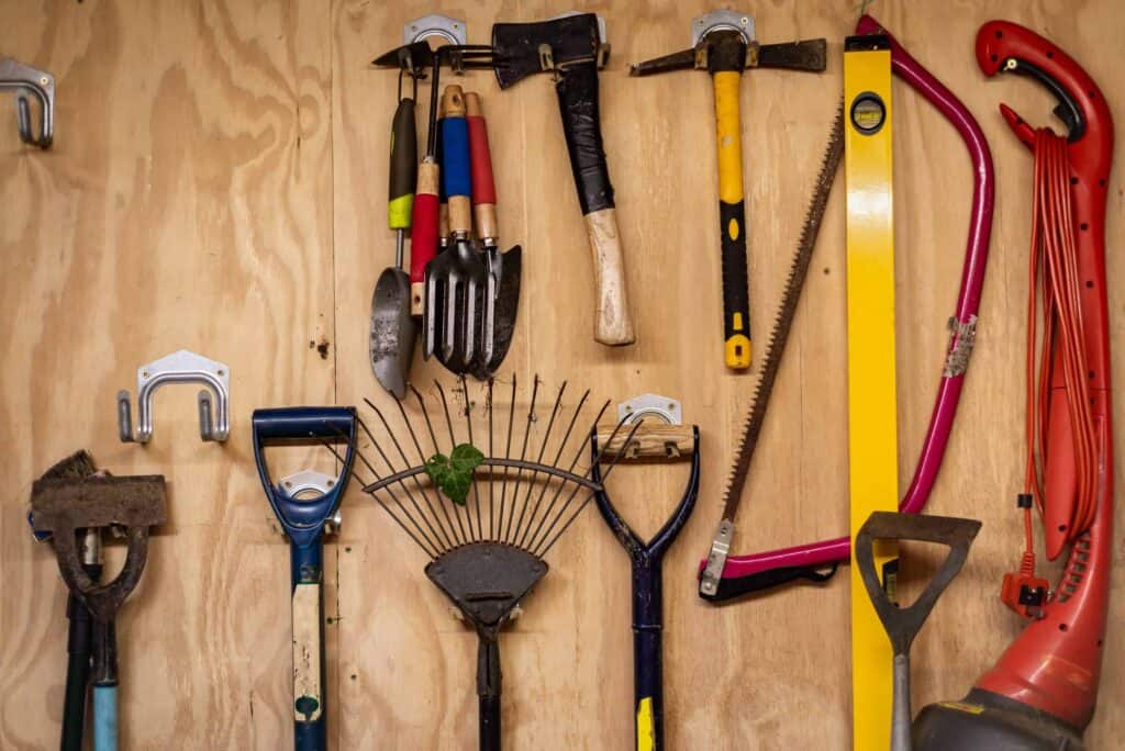 Organized tools hanging on a wooden wall in a shed, including shovels, rakes, a saw, a level, and garden hand tools.