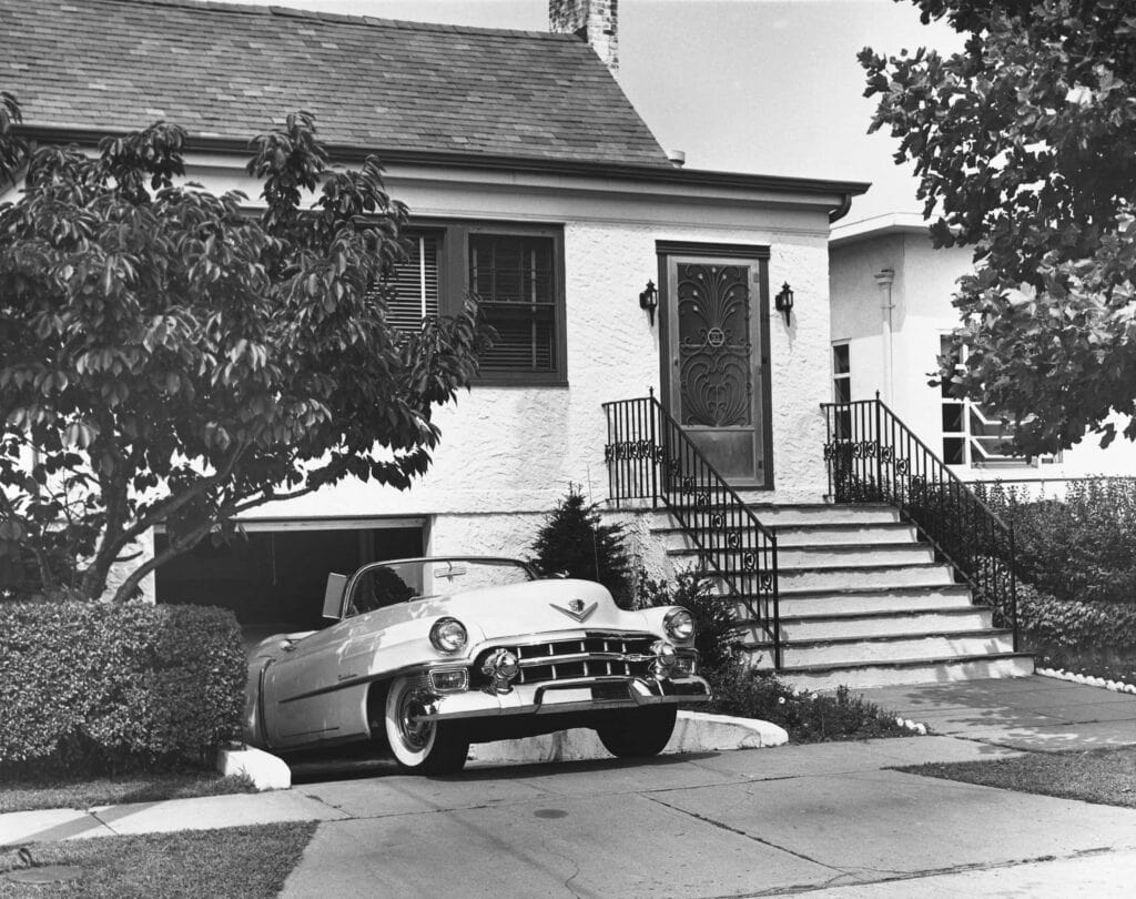 A 1950s house with a car parked partially outside the garage, showing a tree and stairs leading to the front door.