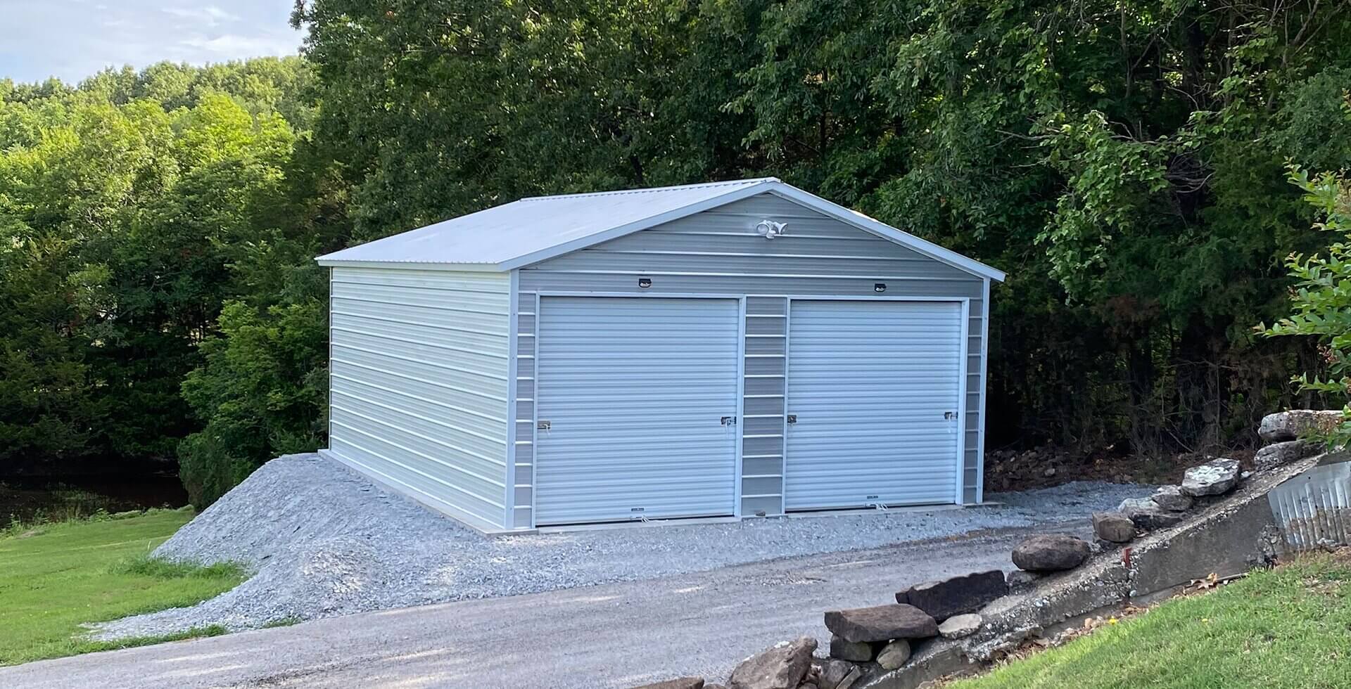 A gray metal garage building with a gabled roof and two white roll-up garage doors, situated on a gravel pad surrounded by dense green trees, with a gravel driveway and landscaping rocks in the foreground.