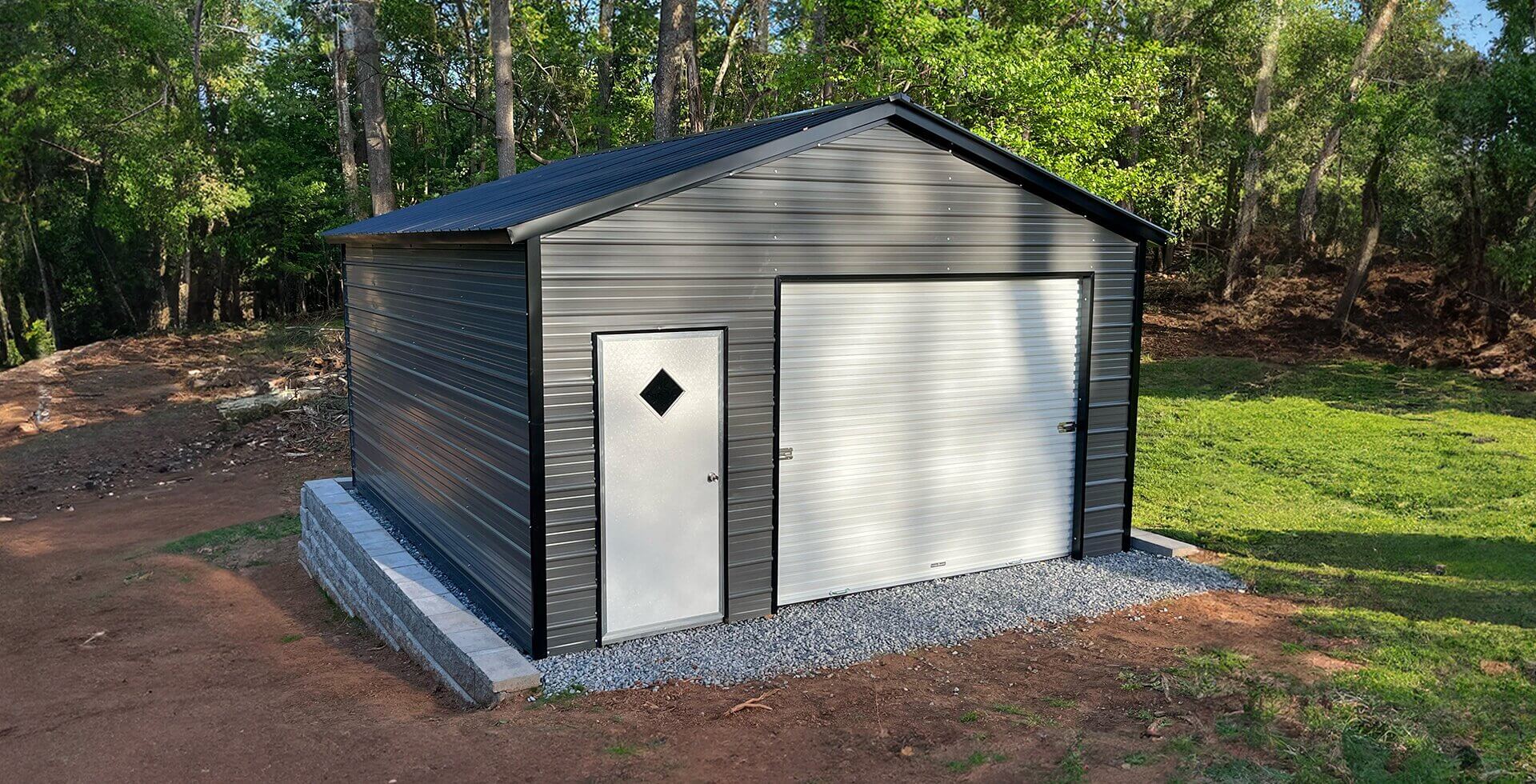 A gray metal garage building with a gabled roof and two white roll-up garage doors, situated on a gravel pad surrounded by dense green trees, with a gravel driveway and landscaping rocks in the foreground.