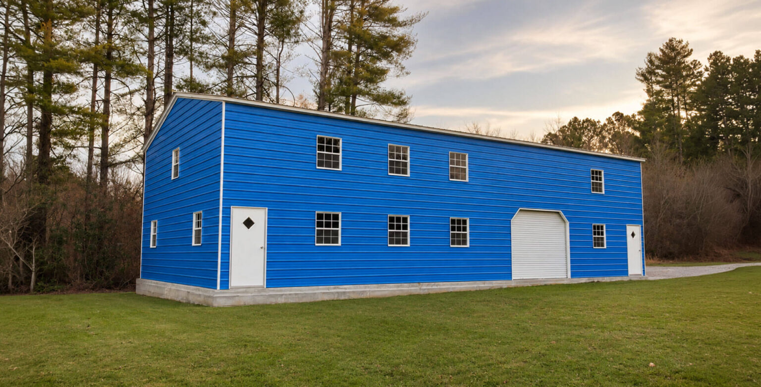 A large two-story blue metal building with white-trimmed windows, a white entry door, and a white roll-up garage door, set on a grassy lawn and backed by tall pine trees under a soft, cloudy sky at dusk.