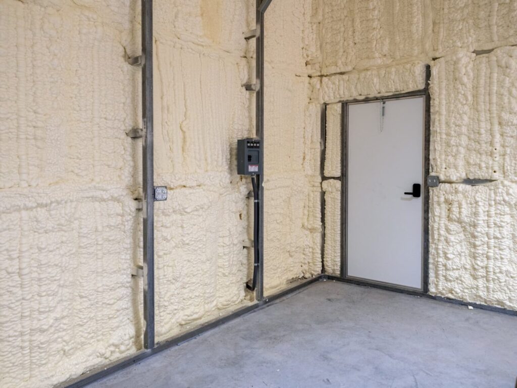 Interior commercial storage unit with closed-cell spray foam insulation on exposed walls, metal framing, electrical panel access, concrete flooring, and insulated utility door for energy-efficient climate control.