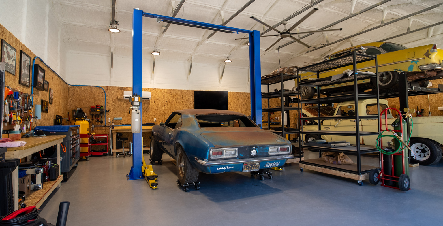 Spacious auto workshop with a blue car lift holding a vintage car, surrounded by tool benches and equipment. Shelving unit stores car parts and another classic vehicle, with gas tanks and tools nearby.