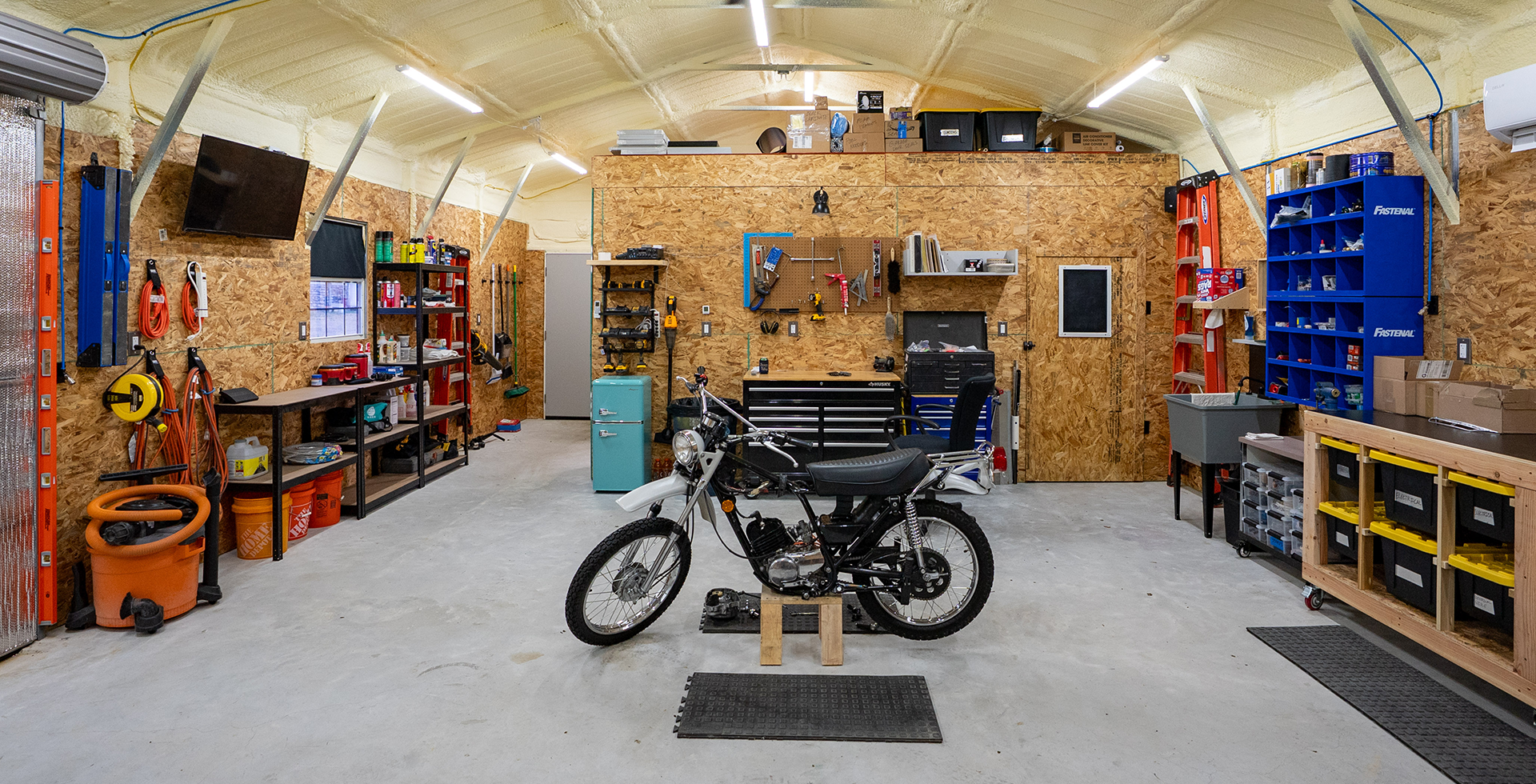 Organized garage workshop with a motorcycle on a stand in the center, surrounded by tool benches and storage shelves. Pegboards, bins, and equipment line the walls under bright overhead lighting and insulated ceiling.