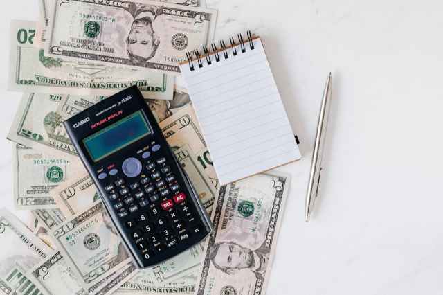 A calculator, a notebook, a pen, and several U.S. dollar bills spread on a white surface, symbolizing cost calculation