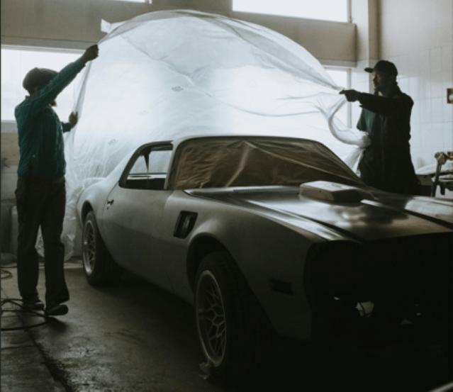 Two people uncovering a classic muscle car inside a garage, with soft lighting filtering through the windows