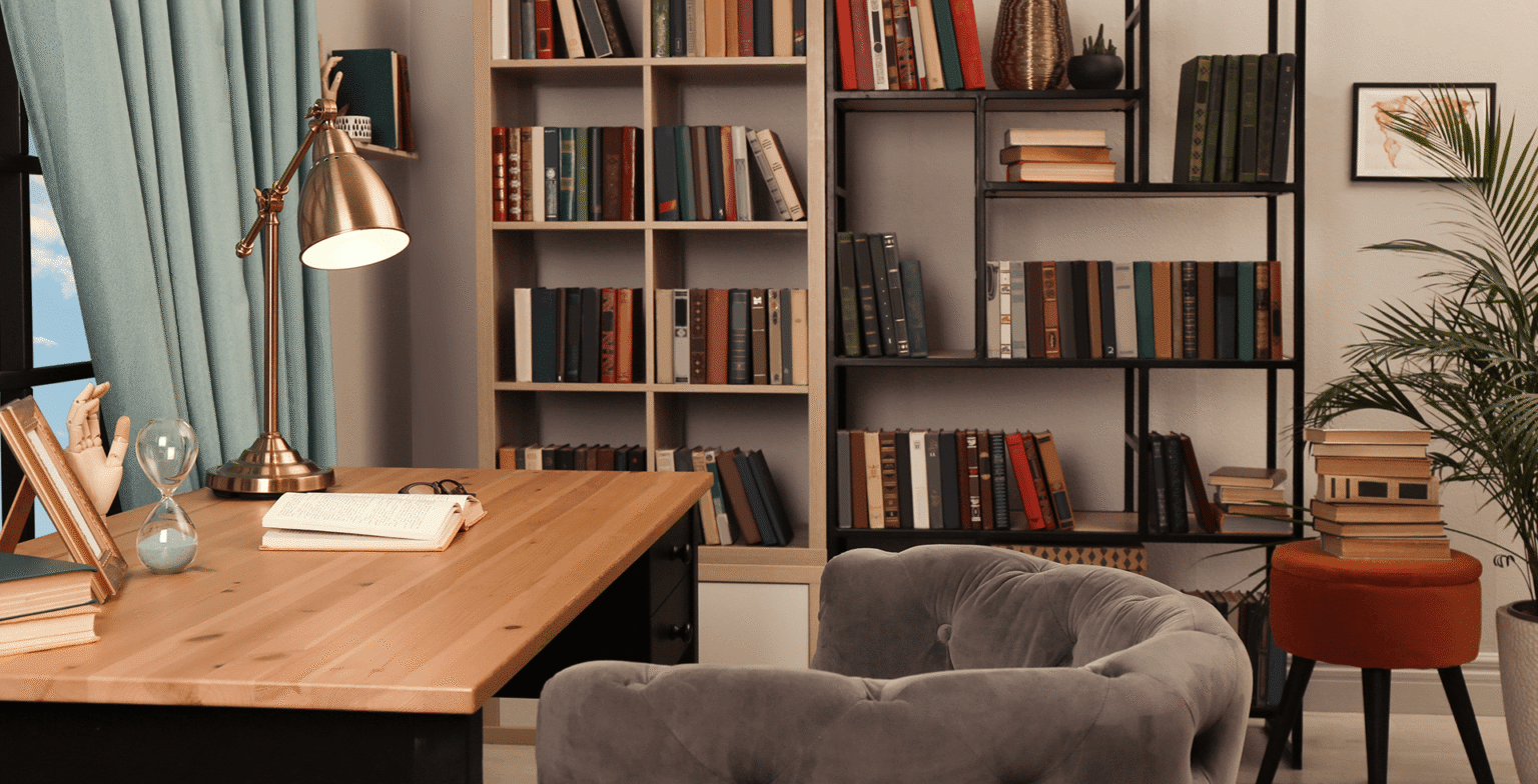 Cozy garage conversion styled as a home office or library, with a wooden desk, desk lamp, upholstered chair, and bookshelves lining the walls.