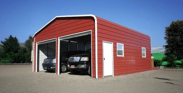 Red metal garage with two open bays, sheltering black SUVs, set on a gravel surface