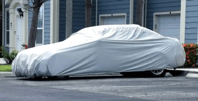 Car parked outdoors and covered with a protective gray car cover in front of a blue residential building