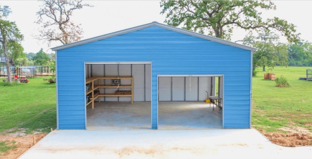 Spacious blue metal garage with two open bays, featuring interior shelving and a workbench, set on a concrete foundation