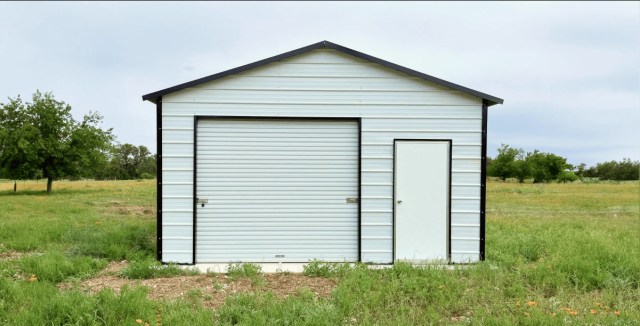 Small white metal garage with a roll-up door and a side entrance, set in an open grassy field