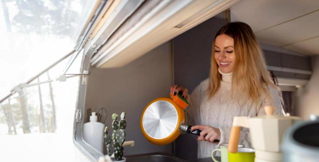 Woman cooking inside an RV kitchen, holding a frying pan near a window with natural light