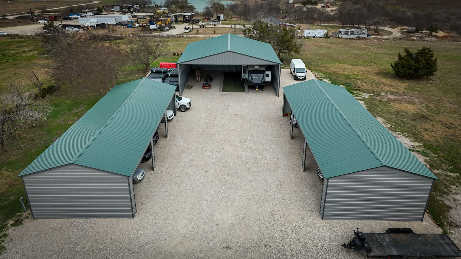 Aerial view of a large metal carport complex with three green-roofed steel structures arranged around a gravel courtyard, providing covered parking for vehicles, trailers, and RVs.