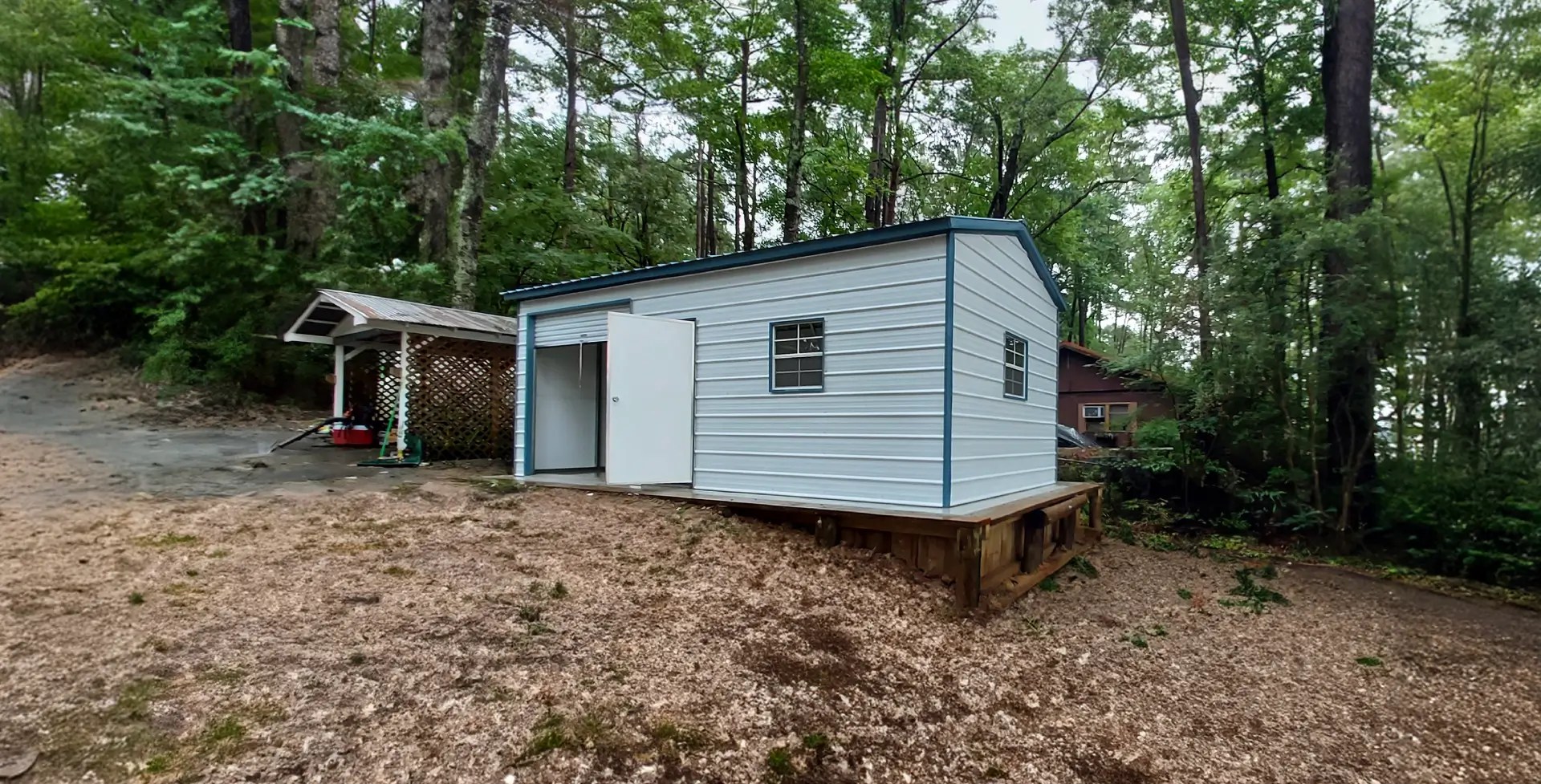 A light blue metal shed with an open door and windows, built on a raised wooden platform in a wooded area.