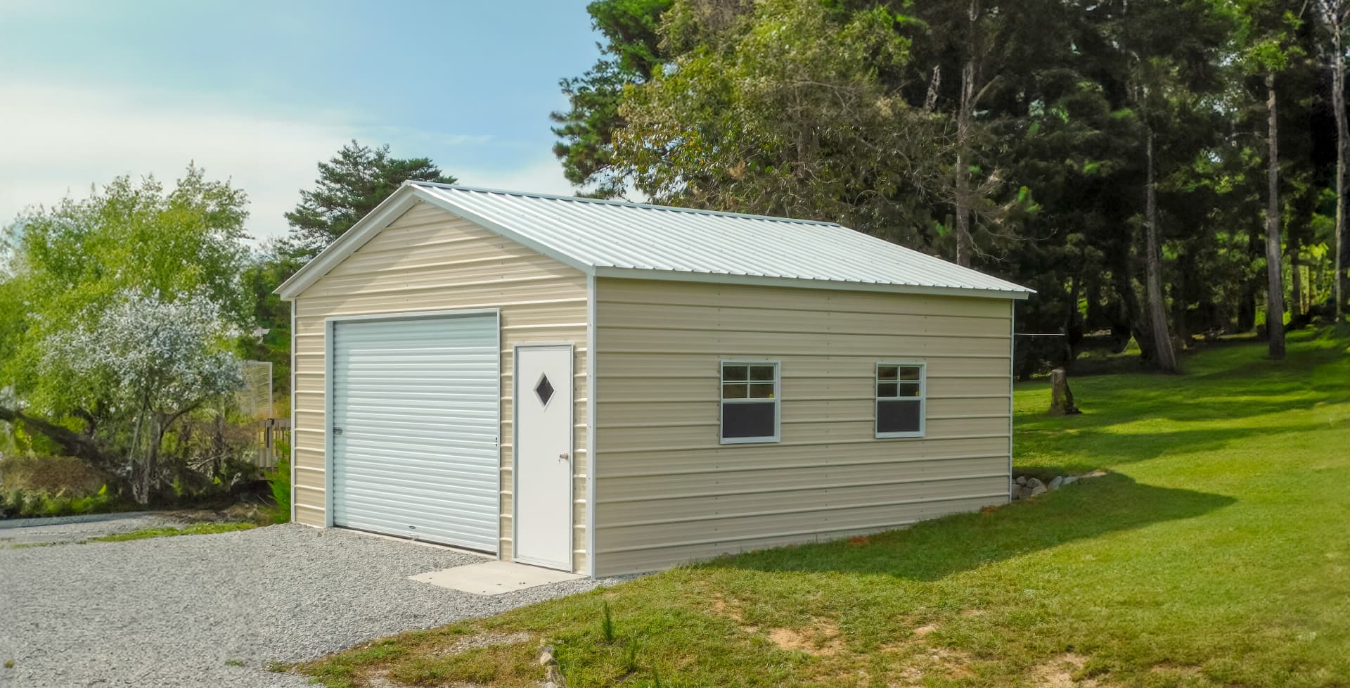 A beige metal garage with a white roll-up door and side entry door, situated on a gravel driveway surrounded by grass and trees.