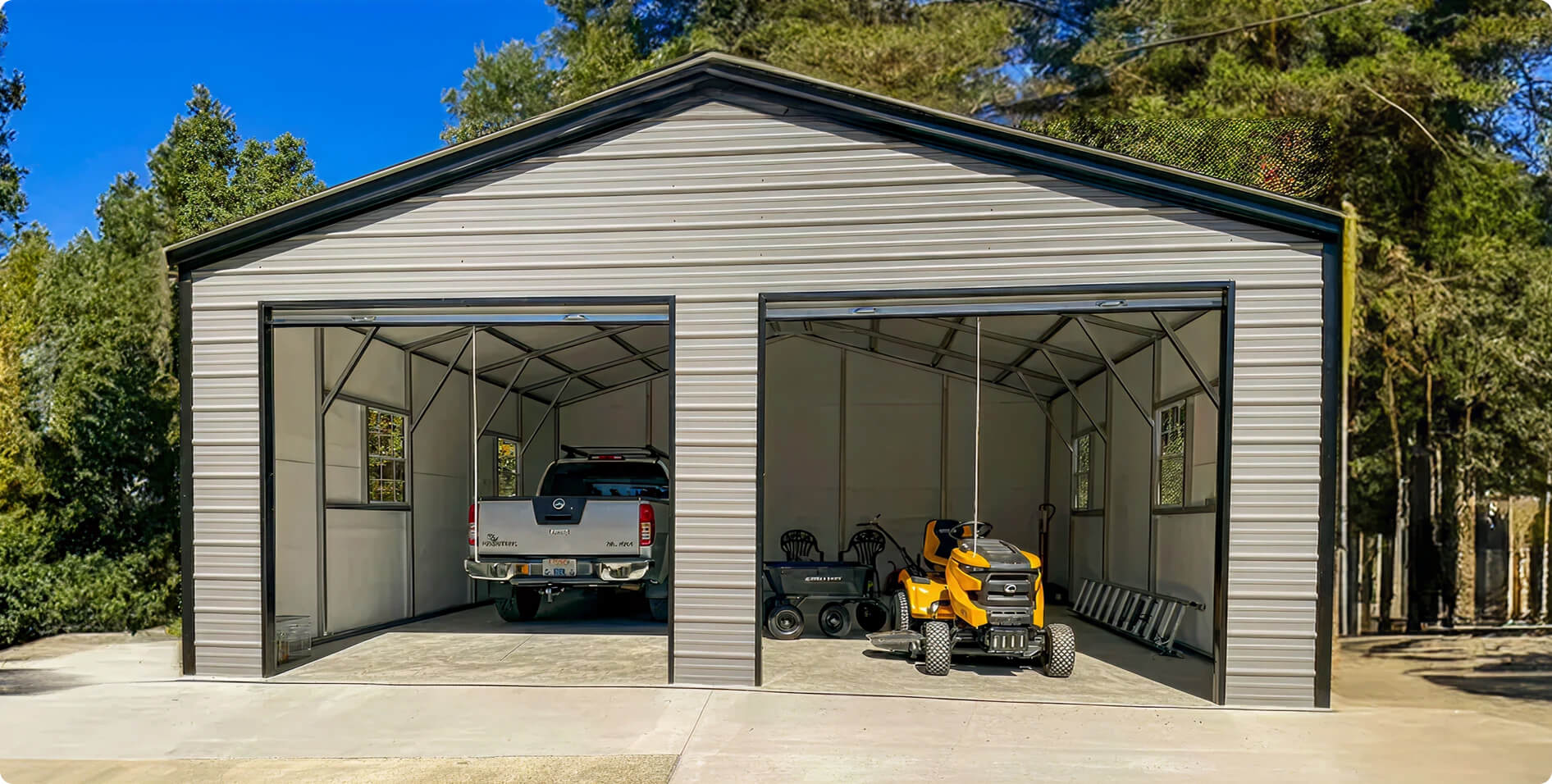 A gray double-door metal garage with a truck parked in one bay and a yellow riding mower in the other, surrounded by trees on a concrete pad.